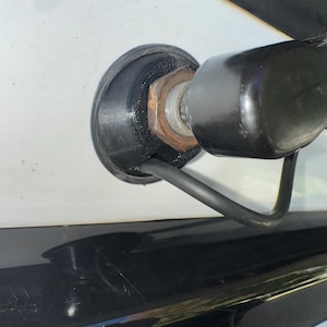 May include: Close-up of a black, rounded component attached to a white surface. A rusty nut and a black cable are visible. The component appears to be part of a larger mechanism.