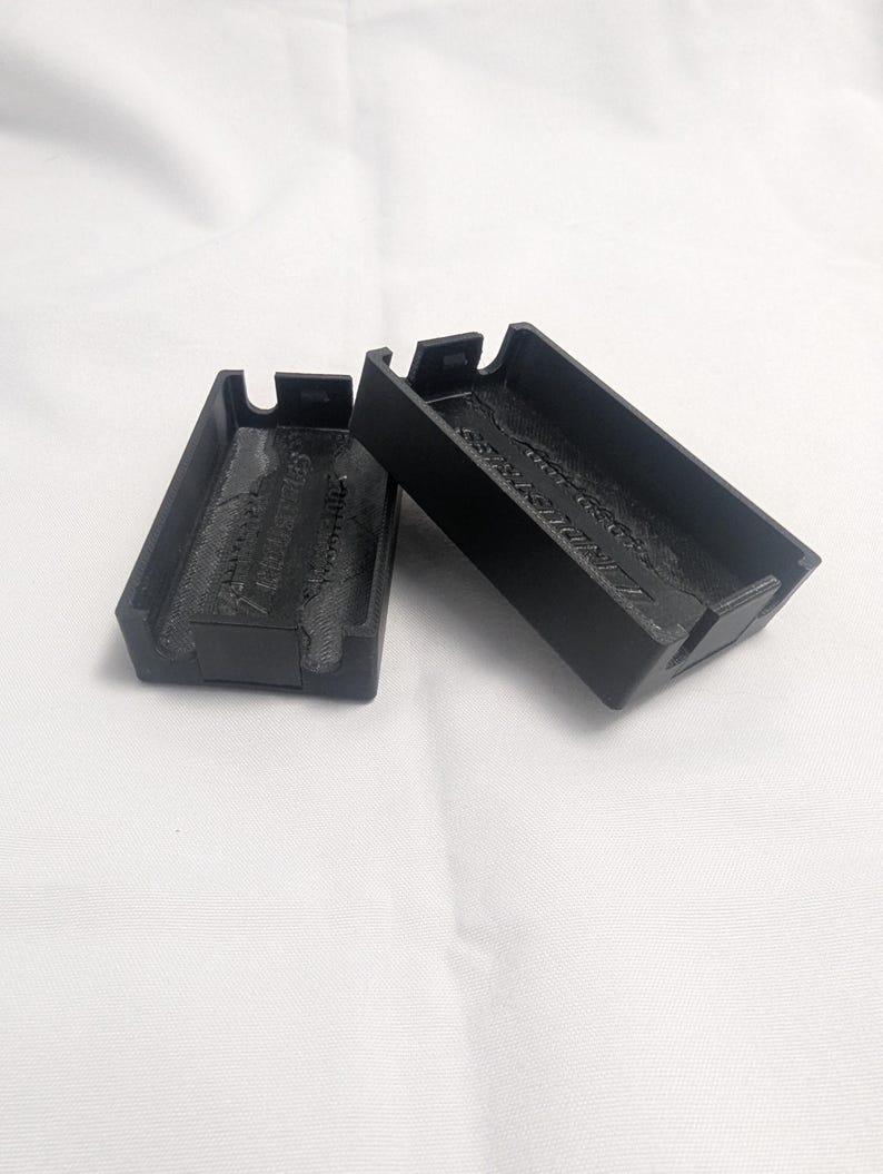 May include: Two black rectangular cases with a textured interior and a small tab on each end. The cases are made of a matte black material and are resting on a white surface. The text "INDUSTRIES" is visible on the interior of one case.