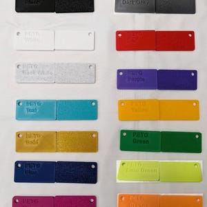 May include: A collection of 14 small plastic squares, each a different color, with the words "PETG" and the color name printed on each square. The colors include black, dark gray, white, red, rock white, purple, teal, yellow, gold, green, blue, lime green, magenta, orange, space gray, and blaze orange.