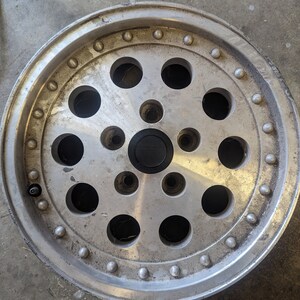 May include: A silver aluminum car wheel with a circular design featuring multiple holes and raised dots around the rim.