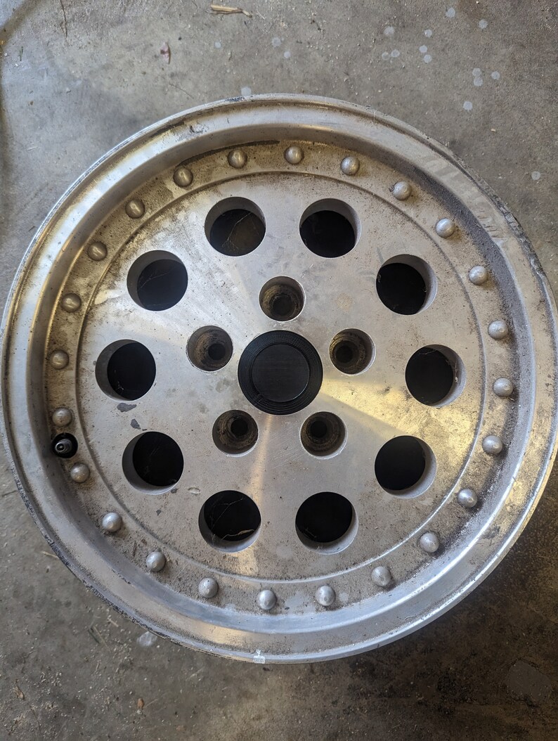 May include: A silver aluminum car wheel with a circular design featuring multiple holes and raised dots around the rim.