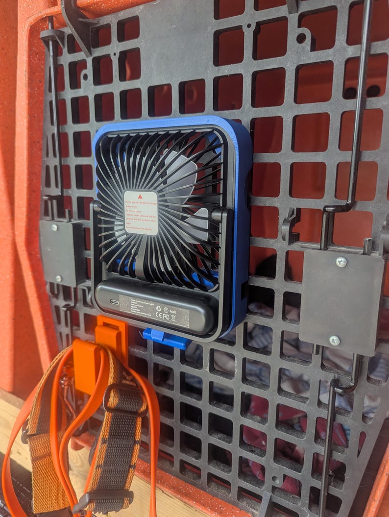 May include: A small, portable blue and black fan mounted on a black grid. The fan has a protective cage and a warning label. An orange strap and a blue clip are also visible.
