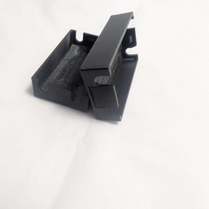 May include: A black, rectangular case with a hinged lid, open to reveal the interior. The case is made of a matte material and sits against a white background. The lid is partially open, showing the inside of the case.