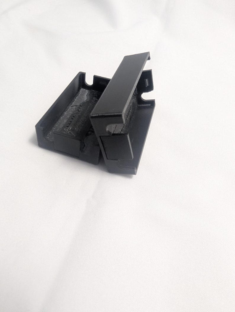 May include: A black, rectangular case with a hinged lid, open to reveal the interior. The case is made of a matte material and sits against a white background. The lid is partially open, showing the inside of the case.