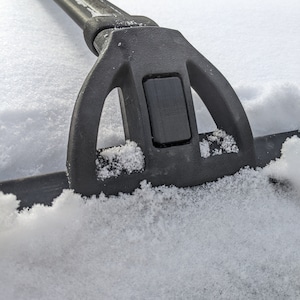 May include: Close-up of a black roof rack crossbar with a snow-covered surface. The crossbar has a textured, rubberized finish and a locking mechanism.