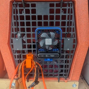 May include: An orange Ruff Land Performance Kennels dog crate with a black grid door. A blue and black fan is attached to the door. An orange strap and a dog harness are hanging from the crate.