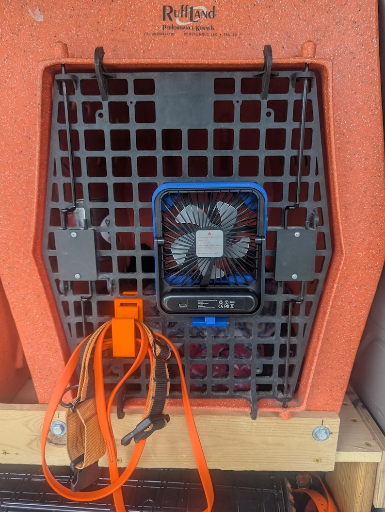 May include: An orange Ruff Land Performance Kennels dog crate with a black grid door. A blue and black fan is attached to the door. An orange strap and a dog harness are hanging from the crate.