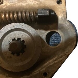 May include: Close-up of a metal gear mechanism with a black plastic knob and a spring. The gear is attached to a larger metal wheel with a central hole.