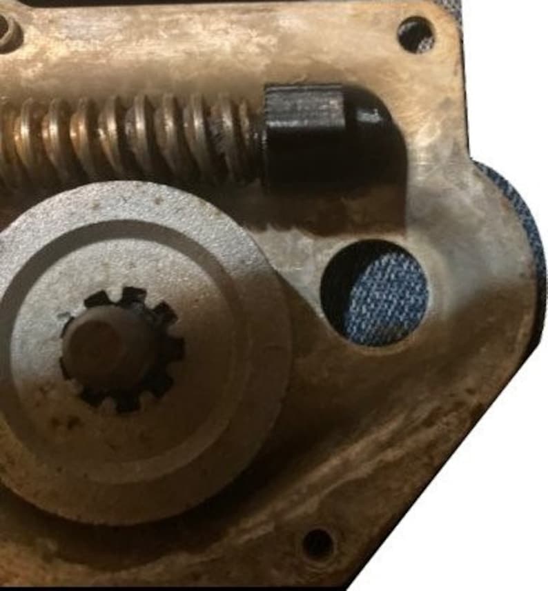 May include: Close-up of a metal gear mechanism with a black plastic knob and a spring. The gear is attached to a larger metal wheel with a central hole.