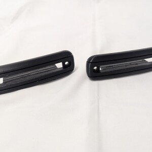 May include: Two black plastic door handles with a rectangular opening in the center. The handles have a textured surface and are designed for use in a car.