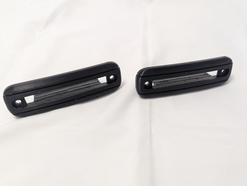 May include: Two black plastic door handles with a rectangular opening in the center. The handles have a textured surface and are designed for use in a car.