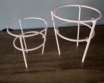 May include: Two white metal plant supports, one larger than the other, with circular frames. The supports are designed to help plants grow upright.