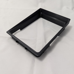 May include: A black plastic frame with a rectangular opening. The frame is made of 3D printed plastic.
