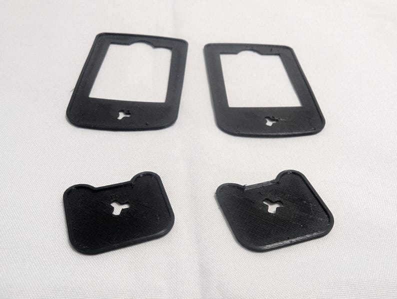 May include: Four black, 3D-printed plastic components. Two are rectangular with a central opening and a small cutout at the bottom. The other two are square with rounded corners and a similar cutout. All are on a white surface.