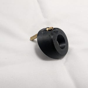 May include: A black, circular object with a brass fitting and a brass rod extending from the side. The object has a hole in the center. The item is set against a white background.