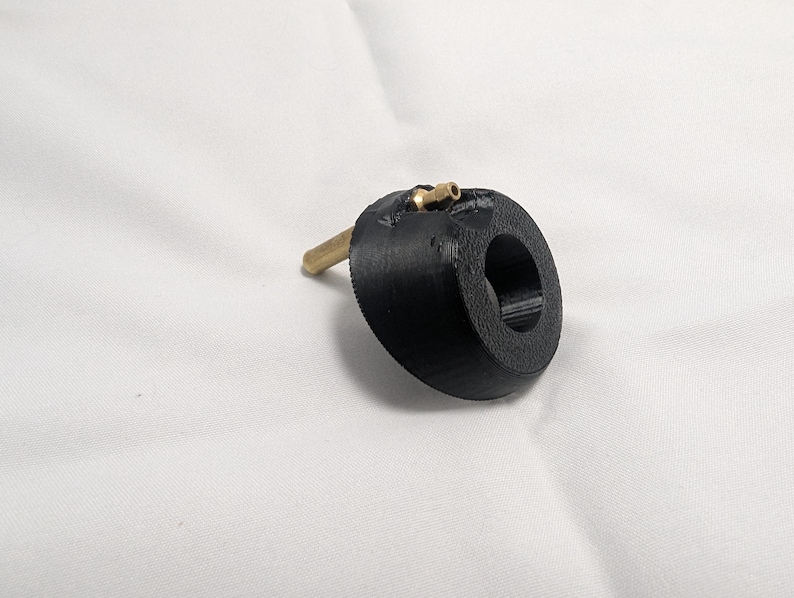 May include: A black, circular object with a brass fitting and a brass rod extending from the side. The object has a hole in the center. The item is set against a white background.