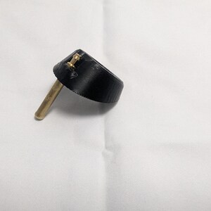 May include: A black bottle stopper with a brass stem and two small brass rivets. The stopper has a conical shape and is designed to fit into the neck of a bottle. The object is set against a white background.