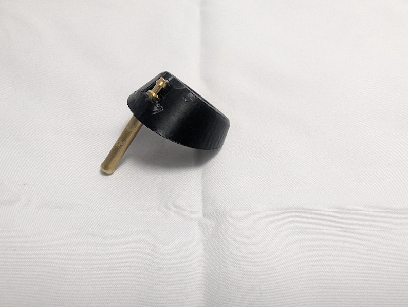 May include: A black bottle stopper with a brass stem and two small brass rivets. The stopper has a conical shape and is designed to fit into the neck of a bottle. The object is set against a white background.