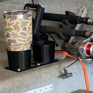 May include: A camouflage-patterned tumbler with a black lid sits in a black cup holder. A second cup holder is empty. A fishing rod holder is mounted above the cup holders. A fishing reel with red line is visible in the foreground.