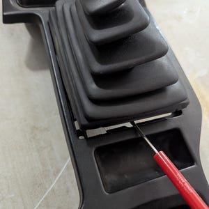May include: A black plastic car interior panel with a black rubber gear shift boot. A red tool is being used to pry the boot off the panel.