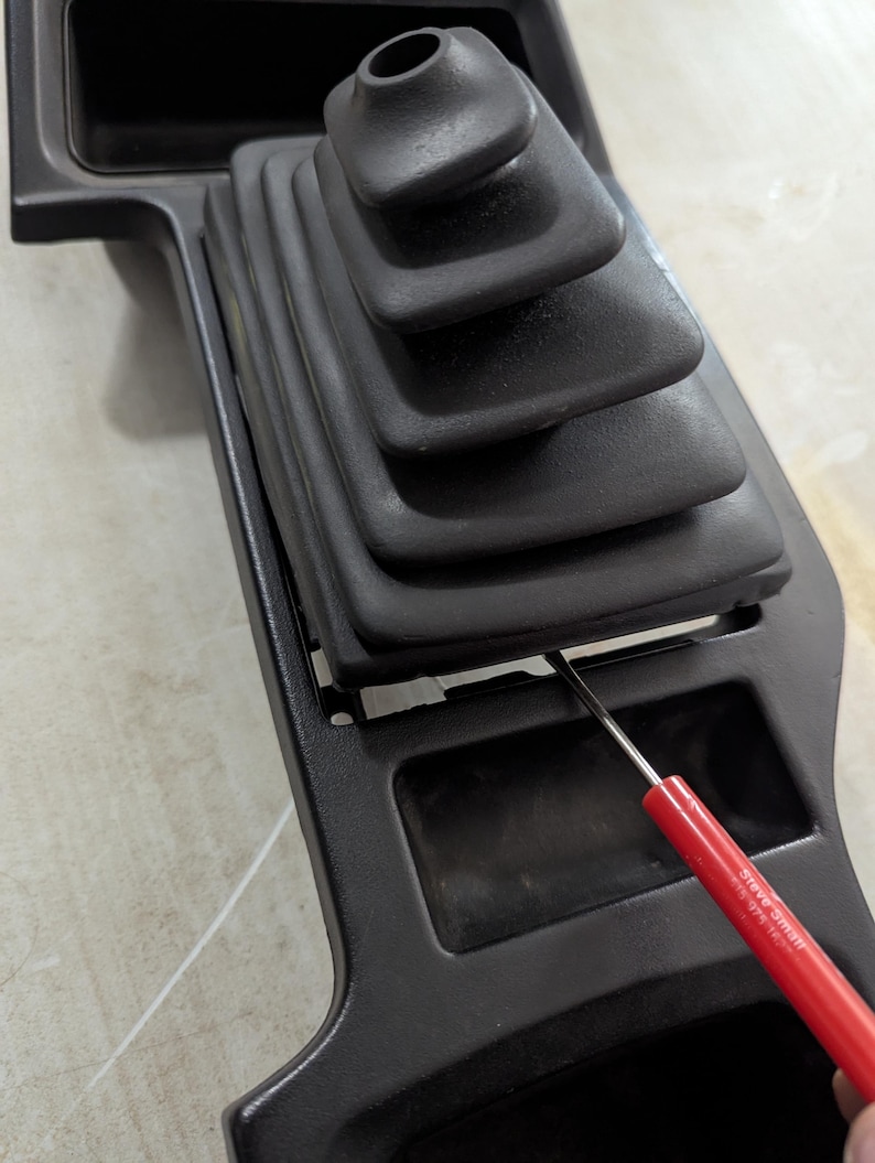 May include: A black plastic car interior panel with a black rubber gear shift boot. A red tool is being used to pry the boot off the panel.
