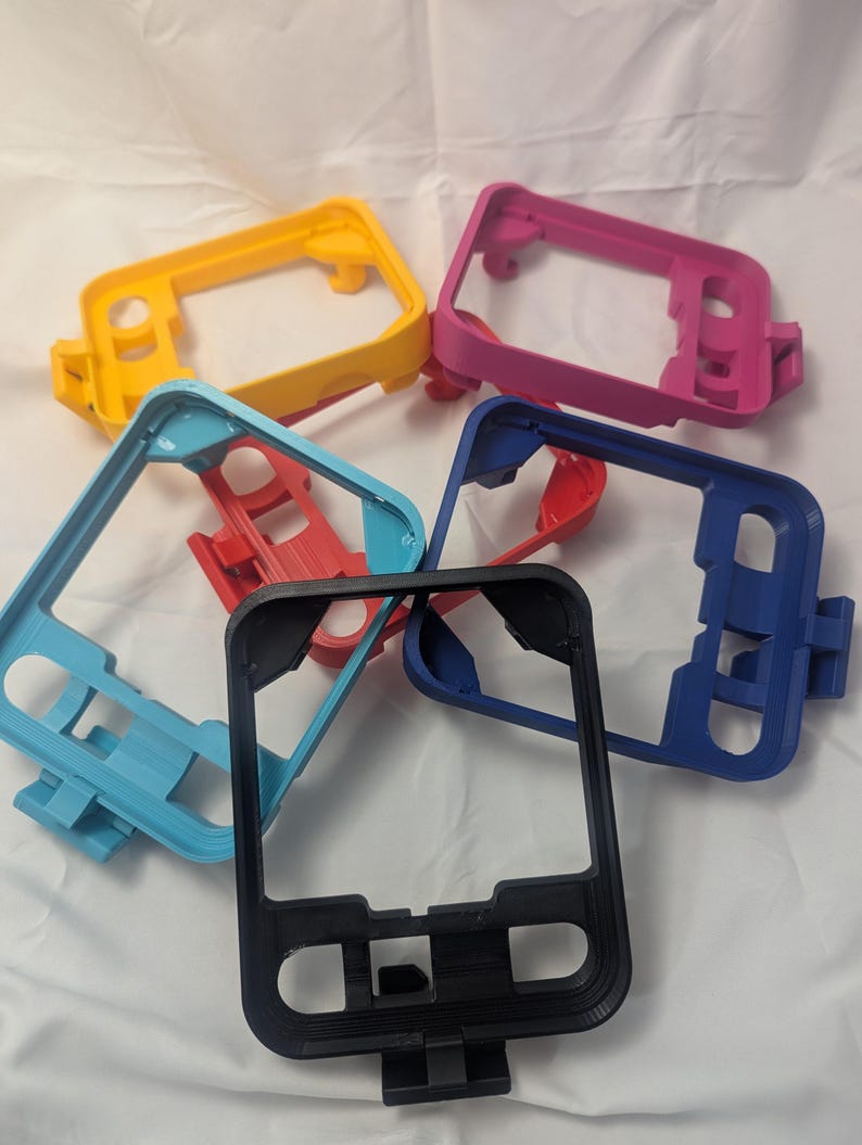 May include: A collection of colorful 3D-printed phone cases in various colors, including yellow, pink, blue, red, and black. The cases have a rectangular shape with cutouts for buttons and a camera.