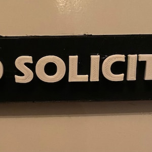 May include: A black rectangular sign with white raised lettering that reads "NO SOLICITING".