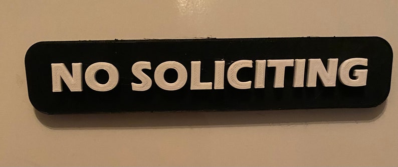 May include: A black rectangular sign with white raised lettering that reads "NO SOLICITING".