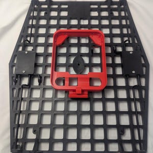 May include: Black, square-patterned panel with a red rectangular frame and black components. The panel has a grid-like design with numerous square openings. The red frame is centered on the panel, and black components are attached to the sides.
