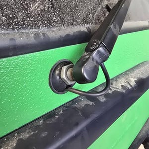 May include: Close-up of a black windshield wiper arm and blade attached to a green vehicle body. The wiper is positioned against a dusty windshield. The vehicle body has a textured green surface with black trim.