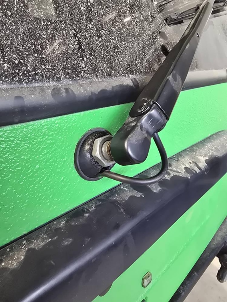 May include: Close-up of a black windshield wiper arm and blade attached to a green vehicle body. The wiper is positioned against a dusty windshield. The vehicle body has a textured green surface with black trim.