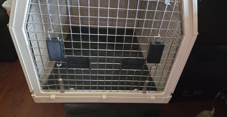 May include: A cream-colored pet carrier with a metal wire door and top. The carrier has a black floor and black latches to secure the door. The carrier is designed for safe pet travel.