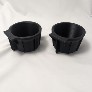 May include: Two black plastic cup holders with a lip on one side. The cup holders are designed to fit into a car's cup holder.