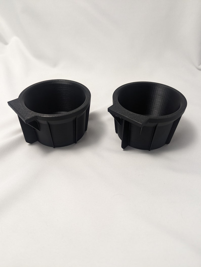 May include: Two black plastic cup holders with a lip on one side. The cup holders are designed to fit into a car's cup holder.