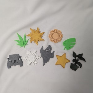 May include: A set of ten 3D printed shapes, including a sun, a ghost, a snowflake, a jeep, a star, a leaf, a marijuana leaf, a plant in a pot, and a mandala. The shapes are made of different colors, including gold, green, white, black, and gray.