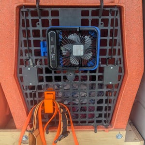 May include: An orange Ruff Land Performance Kennels dog crate with a black plastic grate and a blue and black fan attached. An orange harness and leash are clipped to the crate. The crate is on a wooden platform.