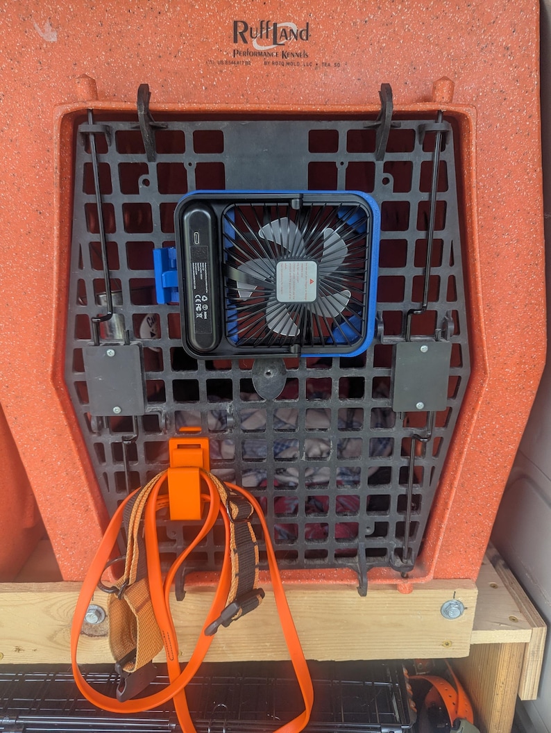 May include: An orange Ruff Land Performance Kennels dog crate with a black plastic grate and a blue and black fan attached. An orange harness and leash are clipped to the crate. The crate is on a wooden platform.