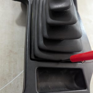May include: Black plastic car interior panel with a black rubber gear shift boot and a small rectangular storage compartment.
