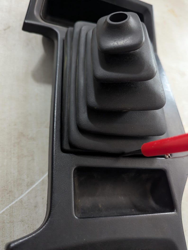 May include: Black plastic car interior panel with a black rubber gear shift boot and a small rectangular storage compartment.