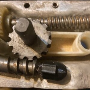 May include: Close-up of a metal gear mechanism with a white plastic lever. The gear is made of metal and has a silver finish. The lever is white and has a smooth surface. The mechanism is part of a larger machine.