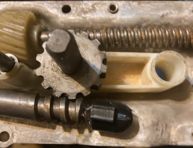 May include: Close-up of a metal gear mechanism with a white plastic lever. The gear is made of metal and has a silver finish. The lever is white and has a smooth surface. The mechanism is part of a larger machine.