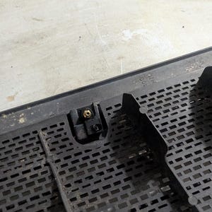 May include: Close-up of a black plastic grille with a small, black plastic latch and a gold-colored screw. The grille has a grid pattern and the text "MADE IN USA" is visible.
