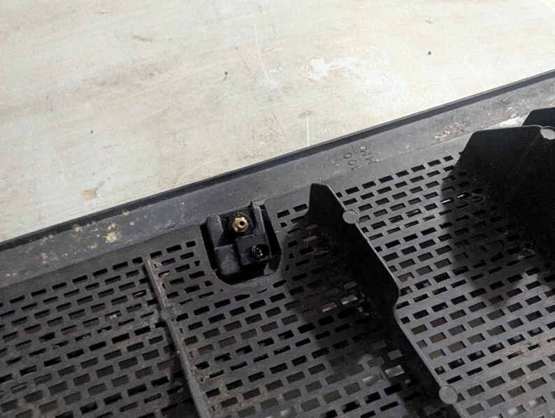May include: Close-up of a black plastic grille with a small, black plastic latch and a gold-colored screw. The grille has a grid pattern and the text "MADE IN USA" is visible.
