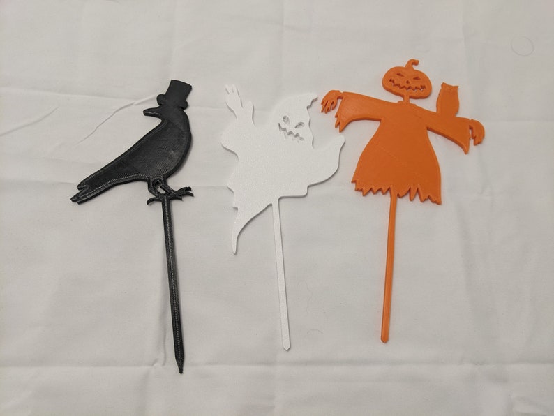 May include: Three Halloween yard decorations: a black crow with a top hat, a white ghost, and an orange scarecrow with a pumpkin head and an owl.