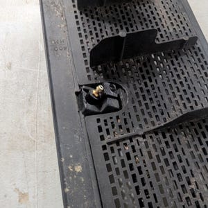 May include: Close-up of a black plastic grate with a small brass fitting. The grate has a grid pattern and the text "NR 007" is visible.