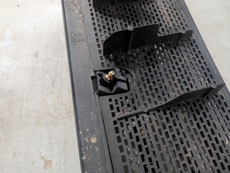 May include: Close-up of a black plastic grate with a small brass fitting. The grate has a grid pattern and the text "NR 007" is visible.
