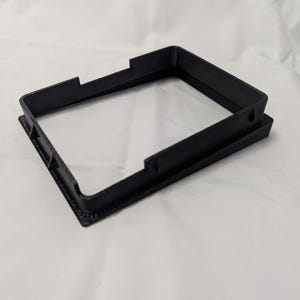 May include: A black plastic frame with a square opening. The frame is designed to hold a small object, such as a picture or a piece of paper.