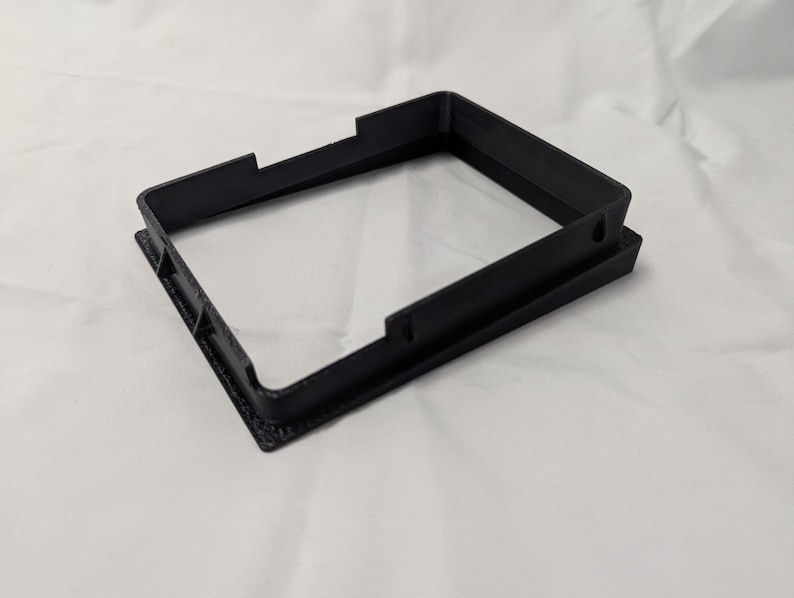 May include: A black plastic frame with a square opening. The frame is designed to hold a small object, such as a picture or a piece of paper.