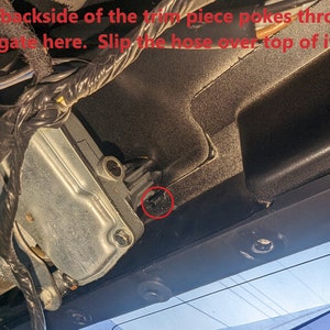 May include: Close-up of a vehicle component with text that reads, "The backside of the trim piece pokes through the gate here. Slip the hose over top of it." The image shows a metal component, wiring, and a black plastic trim.