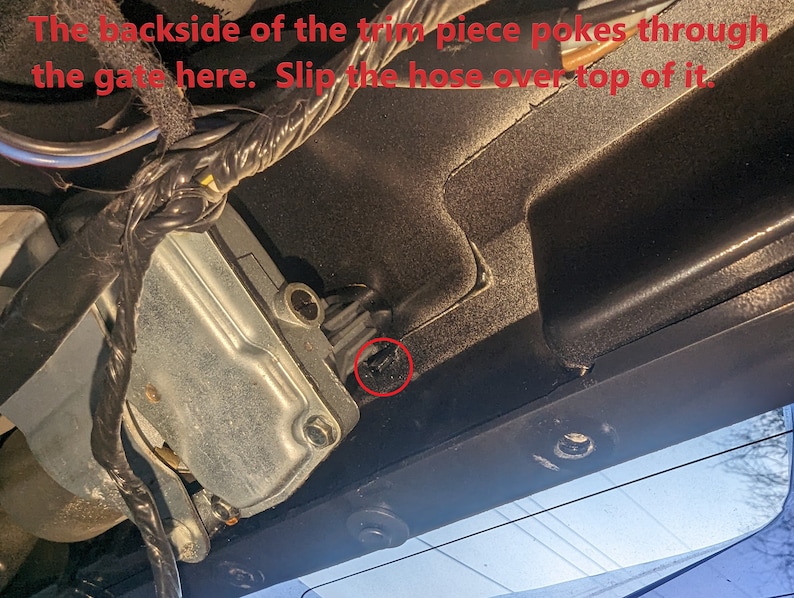 May include: Close-up of a vehicle component with text that reads, "The backside of the trim piece pokes through the gate here. Slip the hose over top of it." The image shows a metal component, wiring, and a black plastic trim.